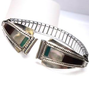 May include: Silver-toned bracelet with an expandable band. The bracelet features two decorative rectangular panels with a dark brown, mother-of-pearl, and green stone inlay. The panels are edged with a twisted rope design.