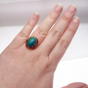 May include: A gold-colored ring with an oval, turquoise-colored stone. The ring has a filigree design around the stone. The ring is on a person's hand.