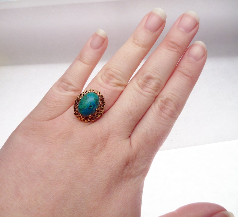 May include: A gold-colored ring with an oval, turquoise-colored stone. The ring has a filigree design around the stone. The ring is on a person's hand.
