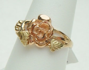 May include: A gold rose ring with leaves. The rose is made of two-toned gold, with the petals being a darker shade of gold than the leaves and band.