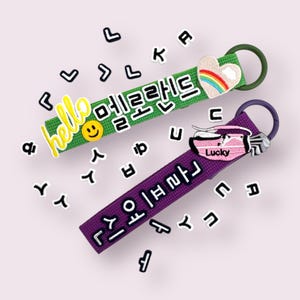 May include: Two colourful keychains with embroidered text and charms. The green keychain has the text "hello 멜로랜드" and a smiley face. The purple keychain has the text "골프여신" and a pink golf bag charm with the word "Lucky".