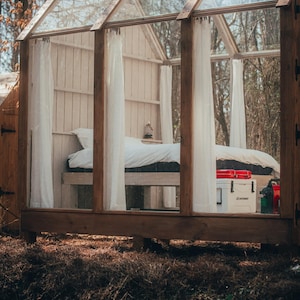 May include: A glass-enclosed cabin with a white bed and white curtains. The cabin is made of wood and has a wooden floor. There is a cooler and other items inside the cabin.