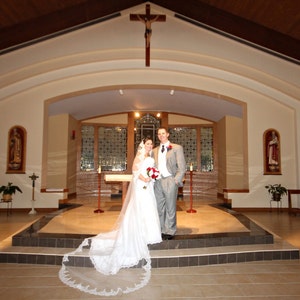 May include: A bride and groom stand in front of an altar in a church. The bride is wearing a white wedding dress with a long veil. The groom is wearing a gray suit and a white shirt. The altar is decorated with flowers and candles.
