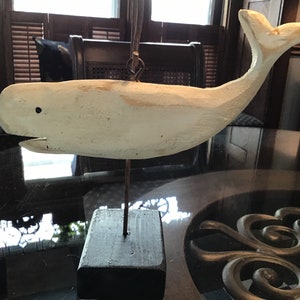 May include: White wooden whale sculpture with a black wooden base. The whale is carved in a simple style and has a small black eye.