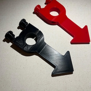 May include: Two 3D-printed slingshots, one red and one black, are displayed against a neutral background. Each slingshot features a circular grip, two prongs for the bands, and an arrow-shaped handle. The red slingshot is on the right, and the black one is on the left.