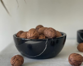 May include: A black marble bowl filled with brown walnuts. The bowl has a white vein running through it. Two walnuts are in the foreground, and more are inside the bowl. The bowl is on a white surface.