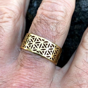 May include: A gold-colored ring with a geometric pattern. The ring is worn on a person's finger.