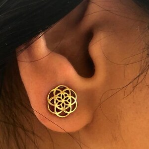 May include: A gold-colored geometric flower-shaped earring. The earring is worn in the left earlobe.