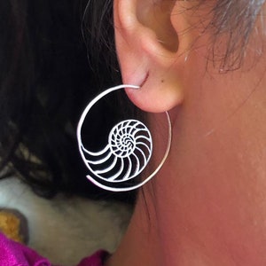 May include: Silver hoop earrings with a nautilus shell design. The earrings are worn in a person's ear.