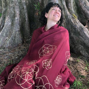 Può includere: Uno scialle bordeaux con un motivo floreale dorato ripetuto. Lo scialle è drappeggiato su una persona seduta vicino a un albero. La persona guarda in alto e sorride. Lo scialle è fatto di un materiale morbido.