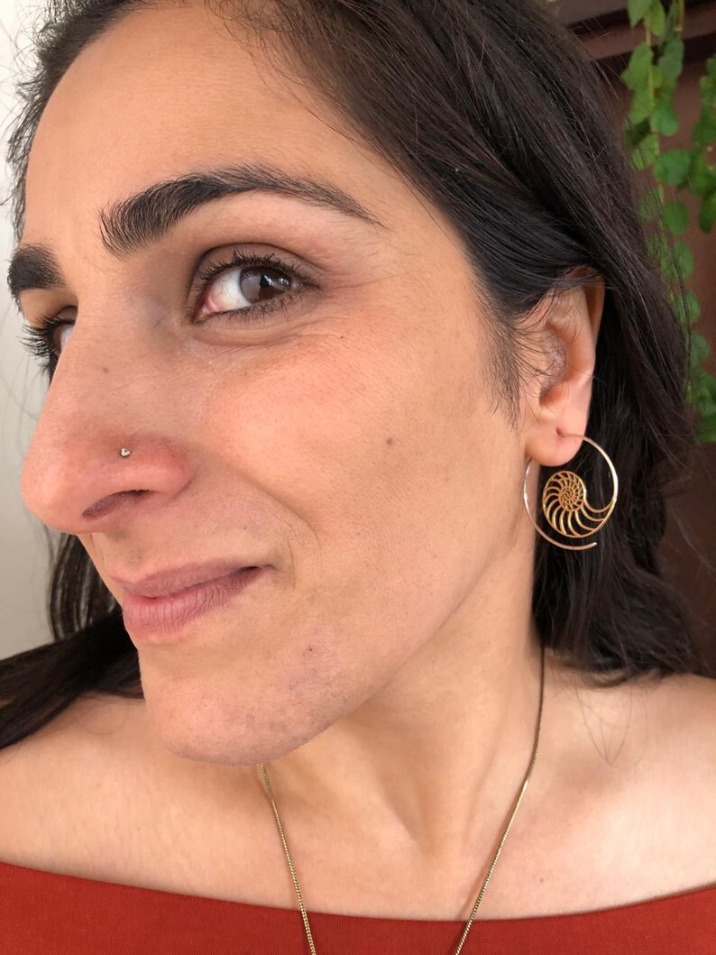 May include: A woman wearing a gold hoop earring with a nautilus shell design. The earring is in her right ear. She is also wearing a small gold nose ring.
