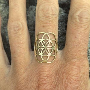 May include: A gold-toned ring with a geometric design, resembling a flower or mandala. The ring is worn on a person's finger.
