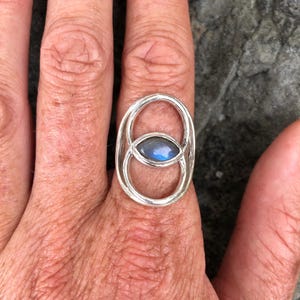 Op de afbeelding: Een zilveren ring met een blauwe, ovale edelsteen in een dubbele cirkelvormig ontwerp.