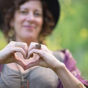 Puede incluir: Una mujer con un sombrero y una camisa de manga larga hace una forma de coraz&oacute;n con sus manos. Lleva varios anillos en sus dedos.