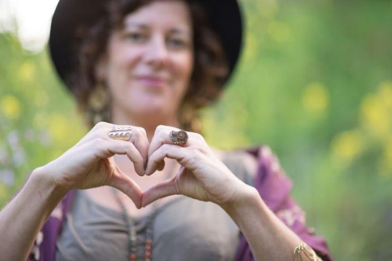 Puede incluir: Una mujer con un sombrero y una camisa de manga larga hace una forma de coraz&oacute;n con sus manos. Lleva varios anillos en sus dedos.