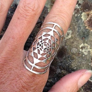 Pode incluir: Um anel de prata com um design geométrico. O anel é usado em um dedo e é feito de um metal que parece ser prata.