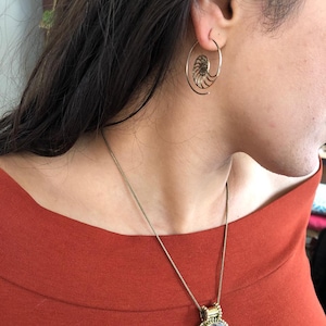 May include: A gold necklace with a large ammonite pendant hangs from a person's neck. The person is wearing a red shirt and has a gold spiral hoop earring in one ear.