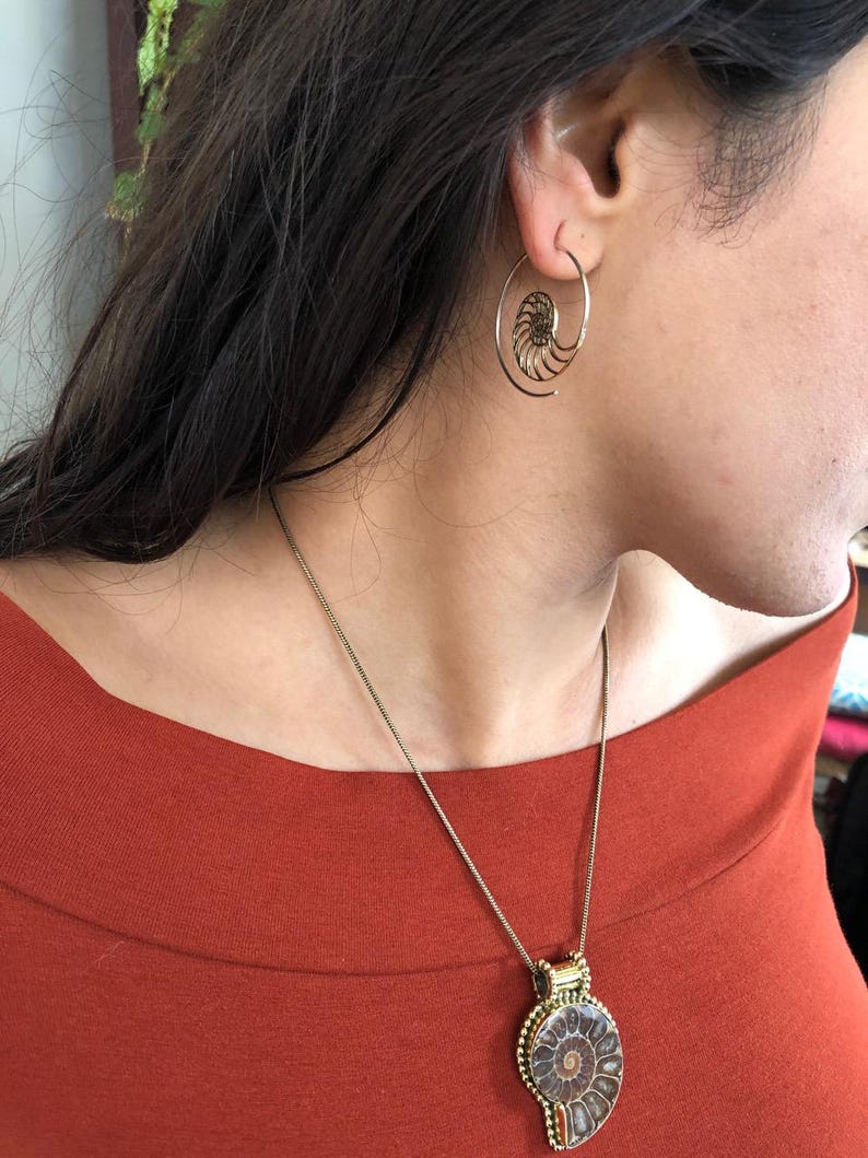 May include: A gold necklace with a large ammonite pendant hangs from a person's neck. The person is wearing a red shirt and has a gold spiral hoop earring in one ear.