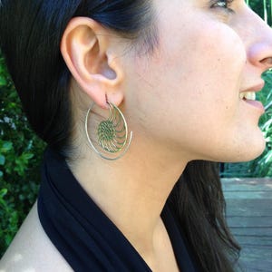 Puede incluir: Un par de pendientes de aro dorados con un dise&ntilde;o en espiral. Los pendientes son usados por una persona con cabello largo y oscuro.