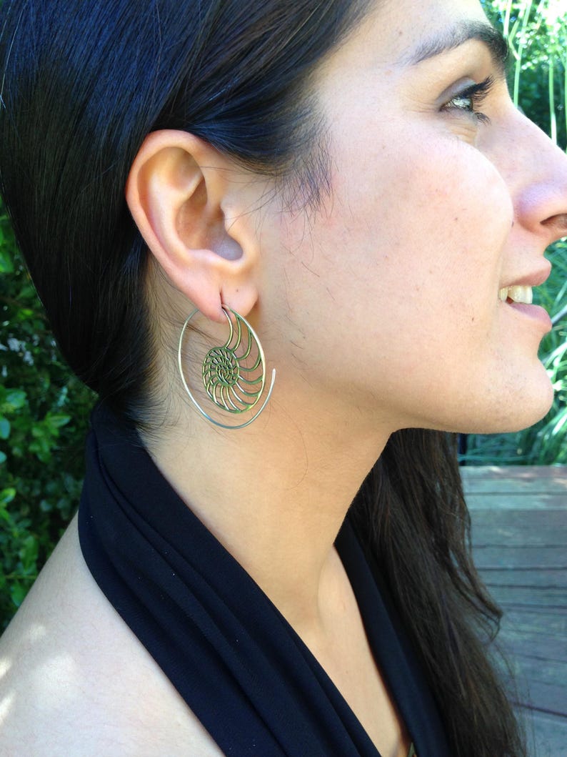 Puede incluir: Un par de pendientes de aro dorados con un dise&ntilde;o en espiral. Los pendientes son usados por una persona con cabello largo y oscuro.