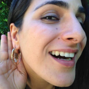 Puede incluir: Una mujer con cabello oscuro sonr&iacute;e y escucha atentamente. Lleva un aro de oro en la oreja izquierda.
