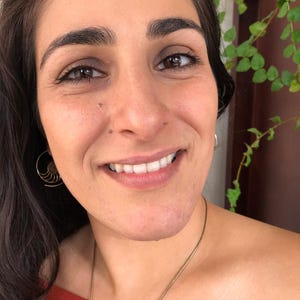 May include: A woman with long dark hair is smiling and wearing a red off-the-shoulder top. She is wearing a gold necklace and gold earrings. She has a nose ring.