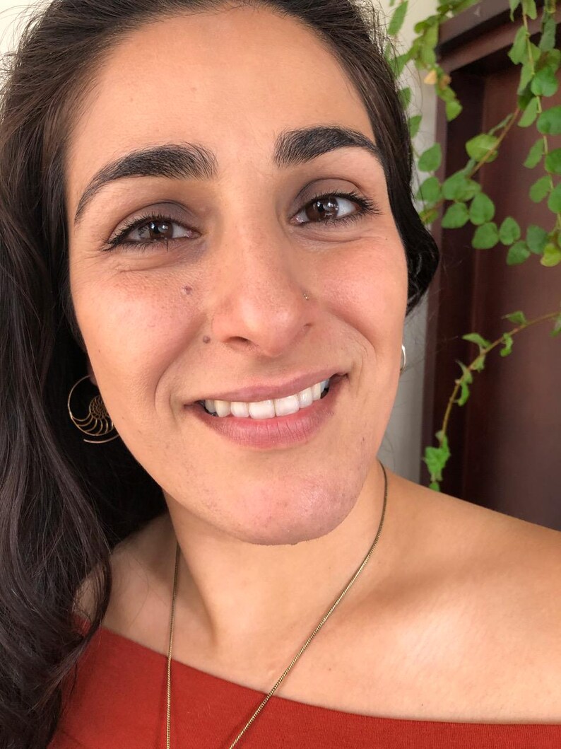 May include: A woman with long dark hair is smiling and wearing a red off-the-shoulder top. She is wearing a gold necklace and gold earrings. She has a nose ring.