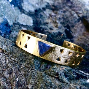 May include: Gold-tone cuff bracelet with a triangular blue stone inlay. The bracelet features a series of triangle cutouts along the band. The jewelry is displayed on a textured, gray and blue stone surface.