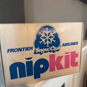 May include: A white box with blue and pink text. The box says "Frontier Snowclub Airlines" and "nipkit".
