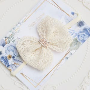 May include: Ivory lace bow headband with a pearl and rhinestone center. The bow is on a cream-colored elastic band. The background features a floral design with blue and white flowers and the text "Petite Dame".
