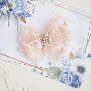 May include: A delicate, light pink lace bow with a pearl and rhinestone center. The bow is set on a card with floral and gold accents. The card has the text "P...Dame" in cursive.