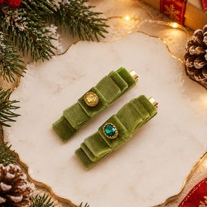 May include: Two olive green velvet hair clips featuring a layered bow design. One clip is adorned with a round, faceted amber-coloured stone, while the other features a round, faceted emerald-green stone, both set in gold-toned metal settings.