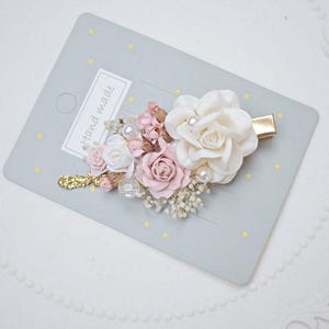 May include: Handmade floral hair clip featuring white and pink roses, pearls, and gold accents. The hair accessory is on a gray card with the words "Hand made". A beautiful hair accessory for special occasions.