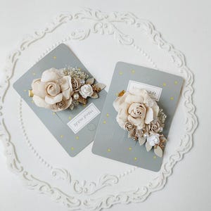 May include: Two handmade floral hair clips. Each clip features a large, light beige rose surrounded by smaller beige and white flowers, gold leaves, and pearls. The clips are on gray cards with gold stars and the text "Hand made".