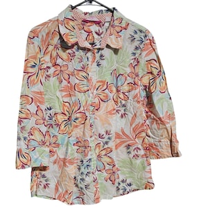 Puede incluir: Una camisa abotonada con estampado floral en tonos naranja, amarillo, rosa y verde. La camisa tiene mangas tres cuartos y cuello. El fondo es blanco.