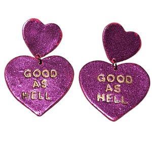 May include: A pair of pink glitter heart-shaped earrings with gold lettering that reads "GOOD AS HELL".