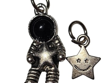 Może przedstawiać: Srebrzysty metalowy charms w kształcie astronauty z czarnym okrągłym wizjerem hełmu oraz oddzielny srebrzysty metalowy charms w kształcie gwiazdki z uśmiechniętą buzią i dwojgiem małych oczu w kształcie gwiazdek, oba z okrągłymi kółeczkami.