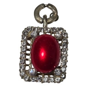 May include: A rectangular pendant with a silver-tone metal frame. The frame is encrusted with clear, round rhinestones. The center features a large, oval, glossy red stone. A silver-tone clasp is at the top.