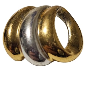 May include: A vintage brooch with a three-loop design. The brooch features two gold-toned loops flanking a silver-toned loop. The loops are smooth and curved, creating a flowing, abstract shape. The brooch is likely made of metal.