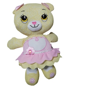 May include: A plush yellow teddy bear with a floral pattern, wearing a pink and yellow dress with a flower accent. The bear has large eyes, a pink heart-shaped nose, and pink bows on its ears. The bear is wearing yellow shoes with flower details.