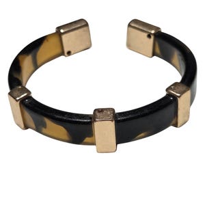 May include: An open cuff bracelet featuring a circular band with a tortoiseshell pattern in shades of black and amber. The bracelet is accented with four rectangular gold-toned metal segments spaced evenly along the band, including two at the ends.