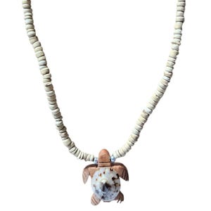 May include: A necklace with a turtle pendant. The pendant is crafted from wood and shell, displaying light brown and white hues. The necklace comprises a strand of small, off-white beads.