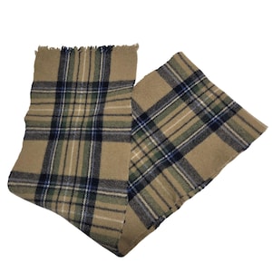 Puede incluir: Bufanda a cuadros beige con borde con flecos. La bufanda presenta un patrón de tartán tradicional en beige, azul marino, verde y blanco. La bufanda está doblada, mostrando el patrón y la textura de la tela.