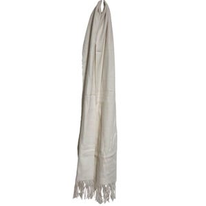 Pode incluir: Um cachecol creme com uma borda com franjas pendura-se sobre um fundo branco. O cachecol é feito de um material macio e fluido e parece leve. A franja está na parte inferior do cachecol.
