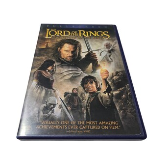 Puede incluir: Una caja de DVD de "El Señor de los Anillos: El Retorno del Rey" con un borde azul. La portada presenta imágenes de personajes de la película, incluido un hombre sosteniendo una espada y un personaje sosteniendo un objeto brillante. El texto incluye una cita.