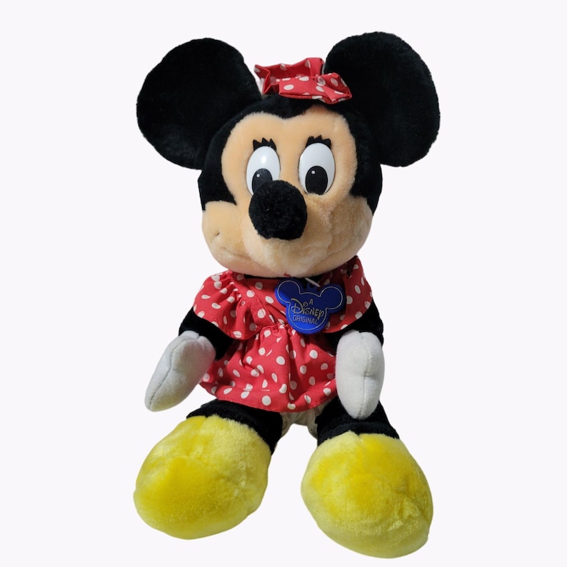 May include: A plush Minnie Mouse toy wearing a red dress with white polka dots and a red bow. The toy has black ears, a black nose, and yellow shoes. The toy has a blue tag that says "Disney Original".