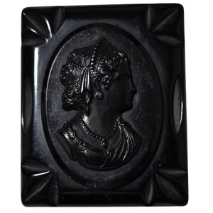 May include: A rectangular black brooch featuring a central oval relief carving of a profile portrait of a person with curly hair, wearing a necklace and draped garment. The frame has decorative leaf-like carvings at each of the four corners.