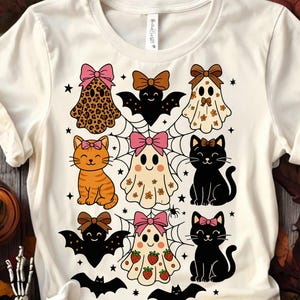 May include: Cream-colored t-shirt featuring a Halloween-themed design. The design includes cartoon ghosts, cats, and bats, each adorned with bows. The ghosts have patterns like leopard print and flowers. The cats are orange and black, and the bats are black.