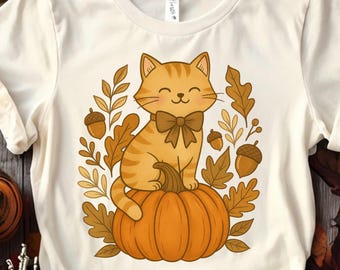 May include: Cream-colored t-shirt featuring a cartoon cat with orange and brown stripes, wearing a bow tie, sitting on an orange pumpkin. The cat is surrounded by fall leaves and acorns, creating a seasonal design.