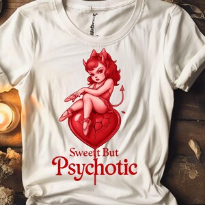 Puede incluir: Camiseta blanca con un gráfico rojo de una mujer diabólica sentada en un corazón roto. La mujer tiene el pelo rojo y lleva un atuendo rojo. El texto "Sweet But Psychotic" está impreso en rojo debajo del gráfico.